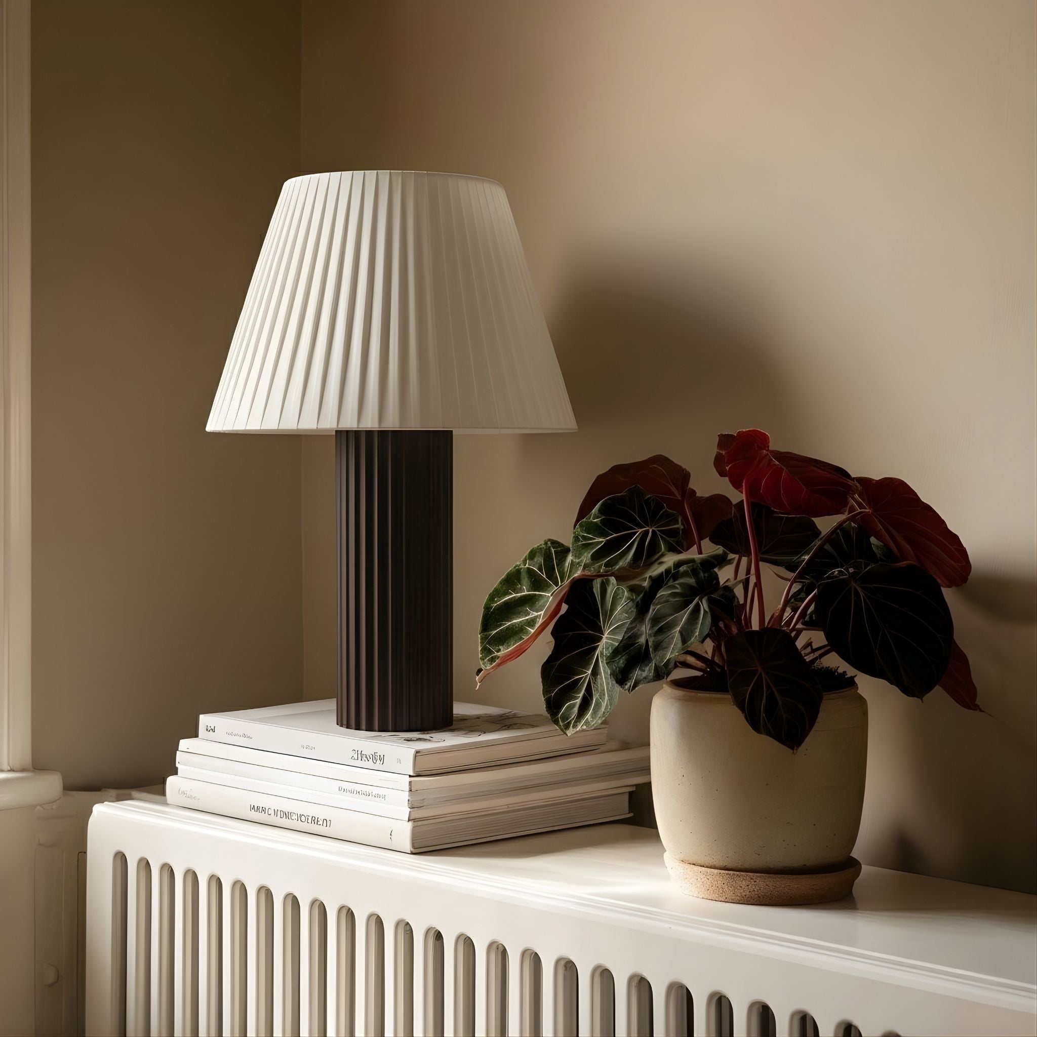 Bordslampa Aristo placerad på element i klassisk inredning med böcker och grön växt, ger ett mjukt och behagligt ljus.