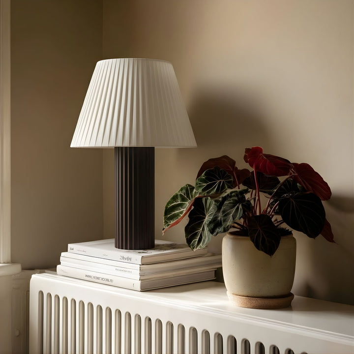 Bordslampa Aristo placerad på element i klassisk inredning med böcker och grön växt, ger ett mjukt och behagligt ljus.