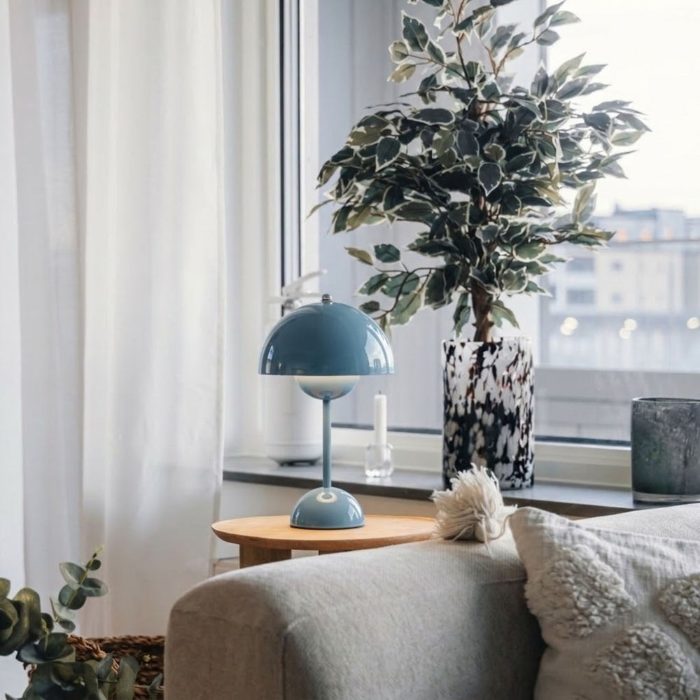 Flowerbud bordslampa i blå finish med rund skärm, placerad på sidobord vid fönster i stilrent vardagsrum med skandinavisk inredning och gröna växter.