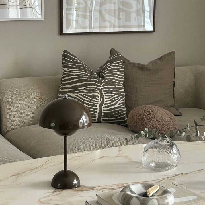 Flowerbud bordslampa i mörkbrun färg med minimalistisk design, placerad på marmorbord framför soffa med dekorativa kuddar.