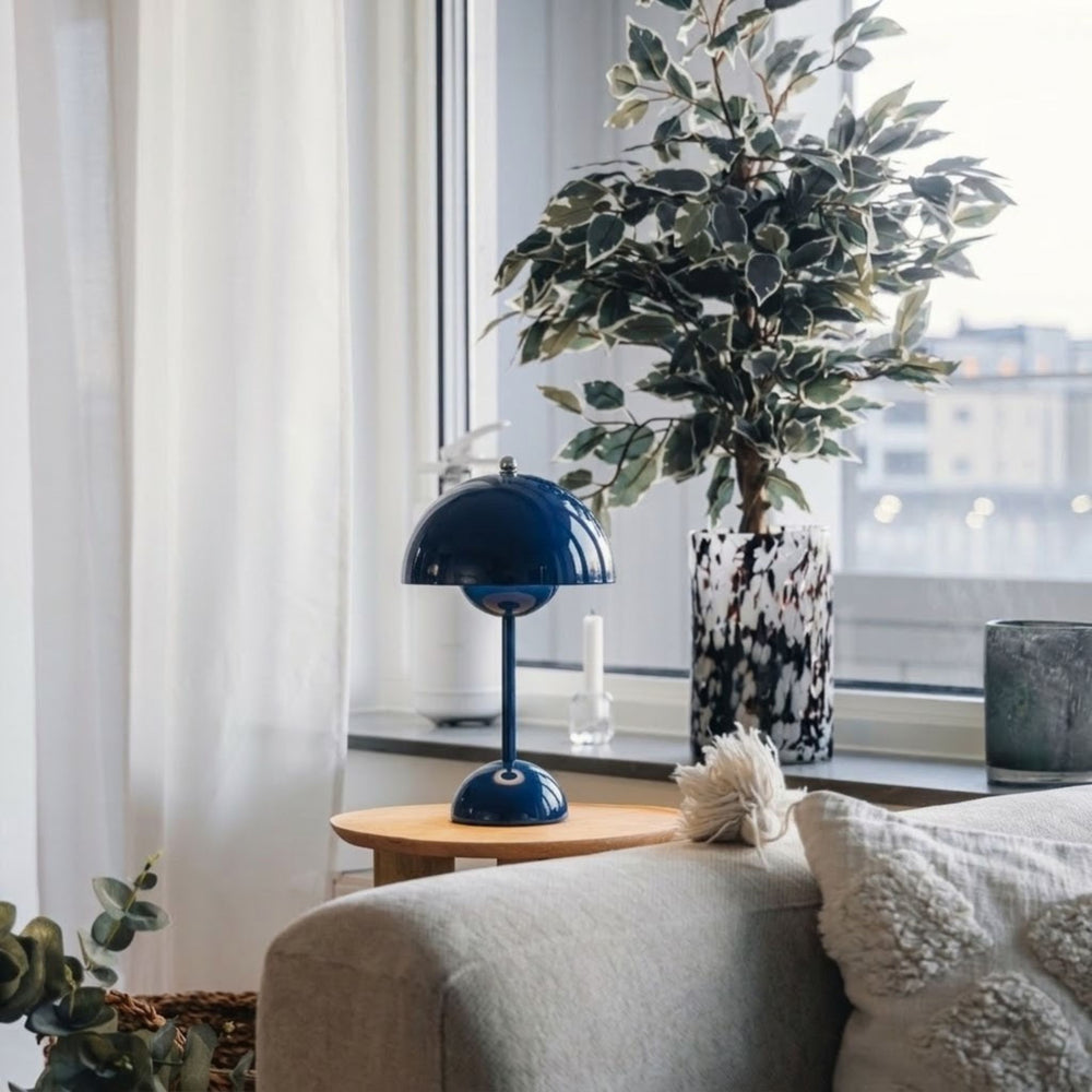 Flowerbud bordslampa i mörkblå finish med rund skärm, placerad på sidobord vid fönster i stilrent vardagsrum med skandinavisk inredning och gröna växter.