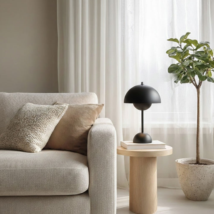 Bordslampa Flowerbud i svart design placerad på runt träbord bredvid beige soffa i ljust skandinaviskt vardagsrum med mjukt naturligt ljus.