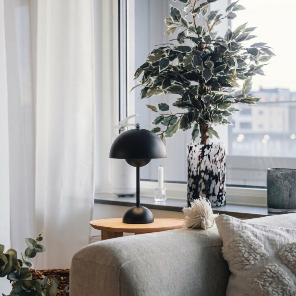 Flowerbud bordslampa i svart finish med rund skärm, placerad på sidobord vid fönster i stilrent vardagsrum med skandinavisk inredning och gröna växter.