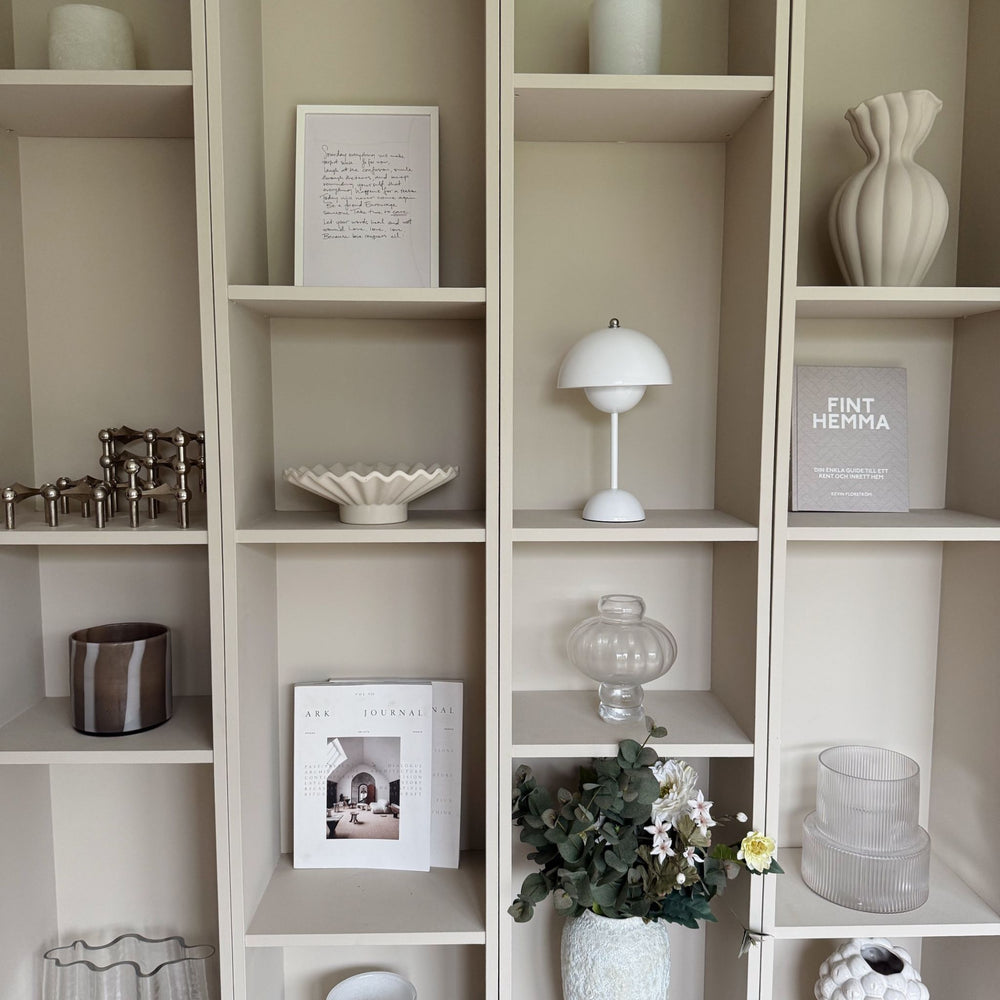 Flowerbud bordslampa i vit färg med rund skärm, placerad i stilren bokhylla med dekorativa objekt i minimalistisk skandinavisk inredning.