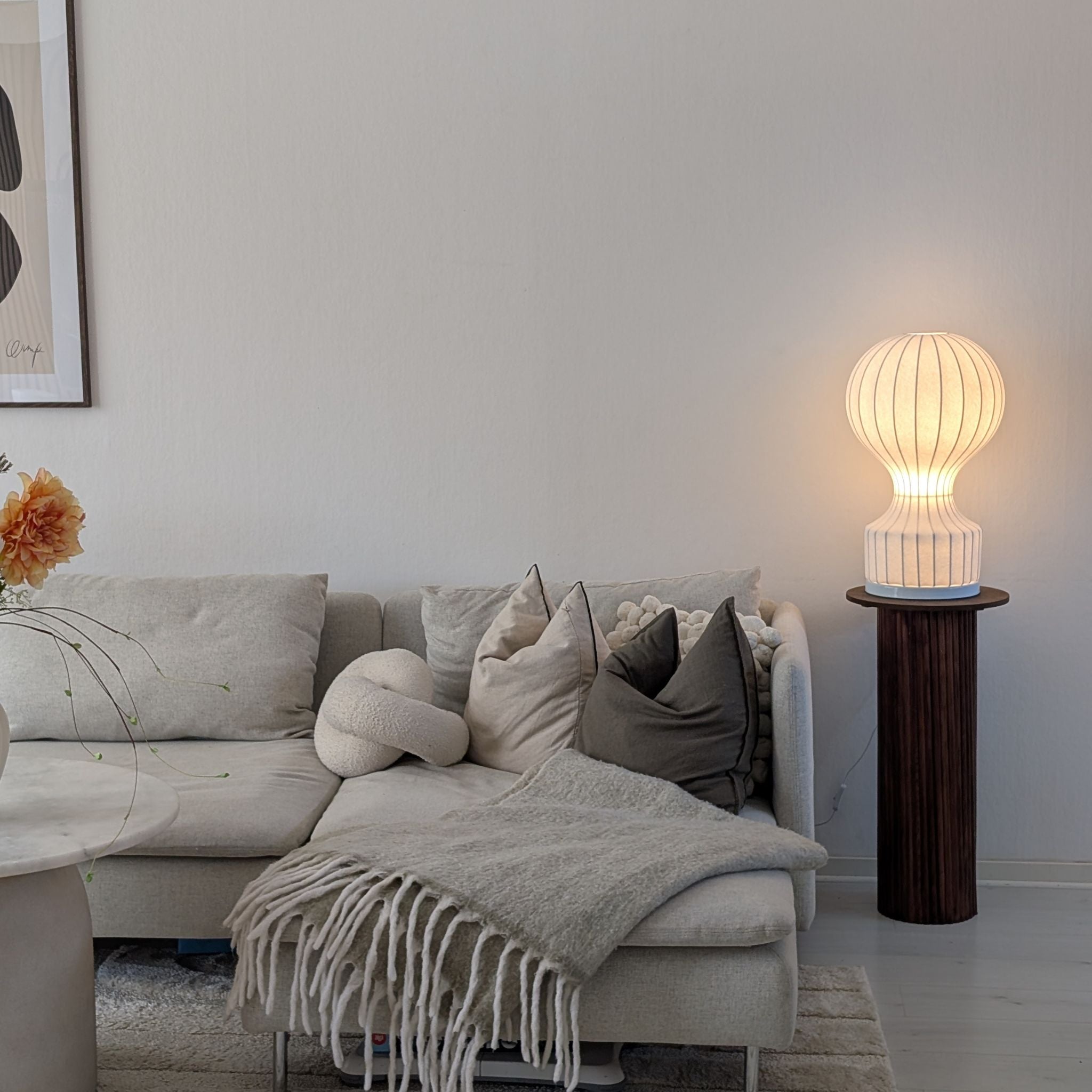 Bordslampa i luftballongsdesign med varm belysning placerad på piedestal bredvid soffa i stilrent skandinaviskt vardagsrum.