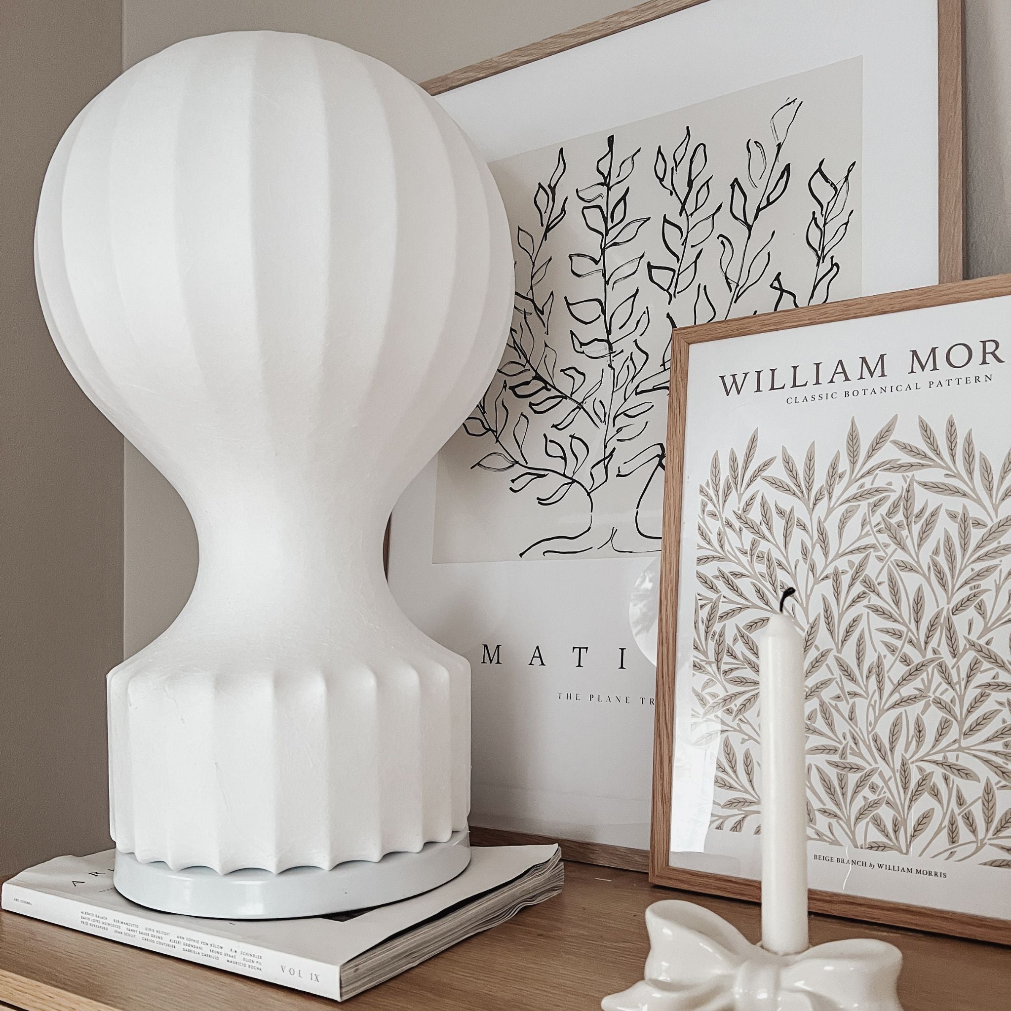 Vit bordslampa i luftballongsdesign med skulptural form, placerad på träbord med stilren inredning och konst i bakgrunden.