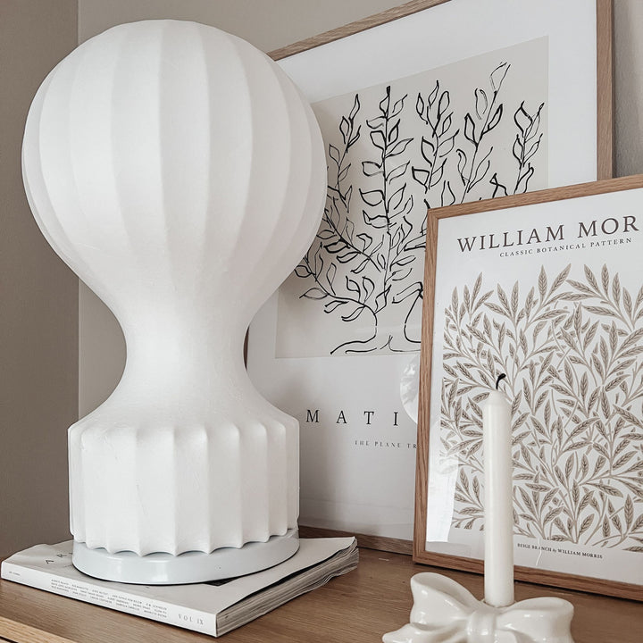 Vit bordslampa i luftballongsdesign med skulptural form, placerad på träbord med stilren inredning och konst i bakgrunden.