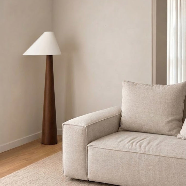 Golvlampa Timber med konformad träbas och vit lampskärm placerad bredvid soffa i ljust skandinaviskt vardagsrum