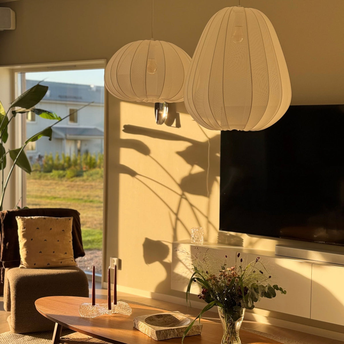 Två vita Sfera taklampor i rund och oval form med mjukt ljus, upphängda i ett skandinaviskt vardagsrum med träbord och varmt kvällssolsken